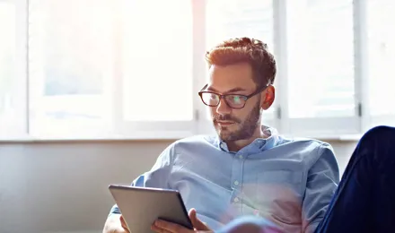 Ein Mann mit Brille sitzt auf einem Sofa und blickt auf ein digitales Tablet