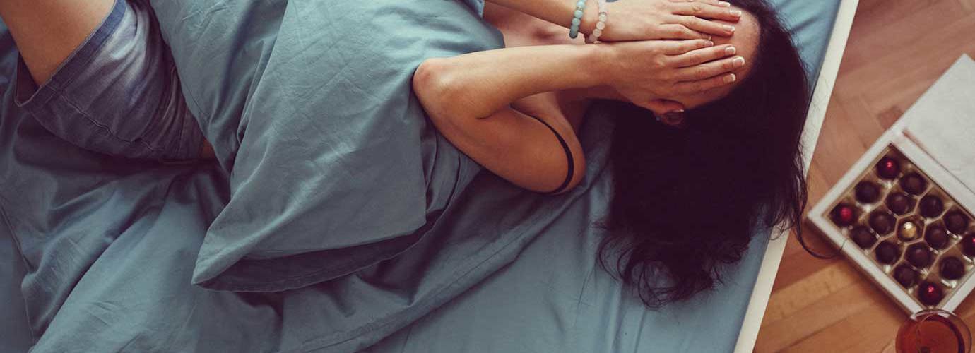 Een vrouw ligt op bed met een kussen op haar buik en haar handen voor haar gezicht