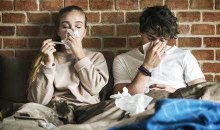 Twee jonge volwassenen zitten onder de dekens te niezen in zakdoekjes met een bakstenen muur in de achtergrond