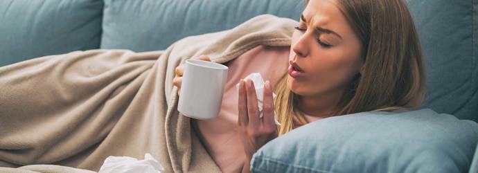 Eine Frau liegt hustend auf dem Sofa und hält eine Teetasse und Taschentücher in den Händen
