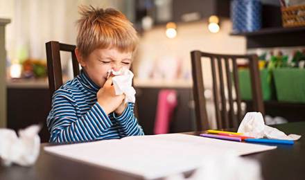 Een kleine jongen snuit zijn neus terwijl hij aan tafel zit met gebruikte tissues om hem heen